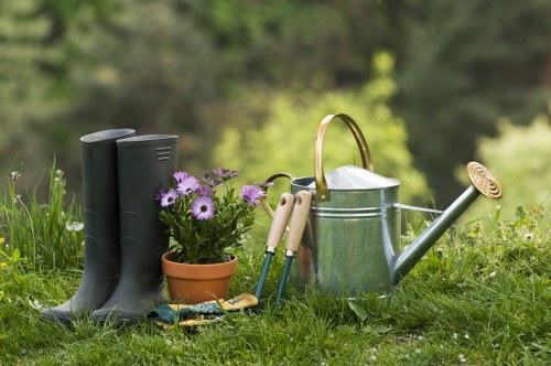 A garden scene in a residential outdoor space featuring a pair of black rubber gardening boots positioned upright on the grass in the foreground, next to a small terracotta flower pot filled with purple and white flowers. Nearby, there are gardening tools with wooden handles, including a pair of pruning shears and a small hand rake, resting on the grass. A metal watering can with a curved spout and a handle is also present, along with a round, ridged garden sieve. The background shows a blurred view of trees and green foliage, suggesting a lush, well-maintained garden area typical of Tooting, with natural daylight illuminating the scene, indicating fair weather. The overall setting highlights outdoor gardening activities and the essential tools used for garden care, aligning with professional gardening and landscaping services offered by Hedge Trimming Tooting in the local area.