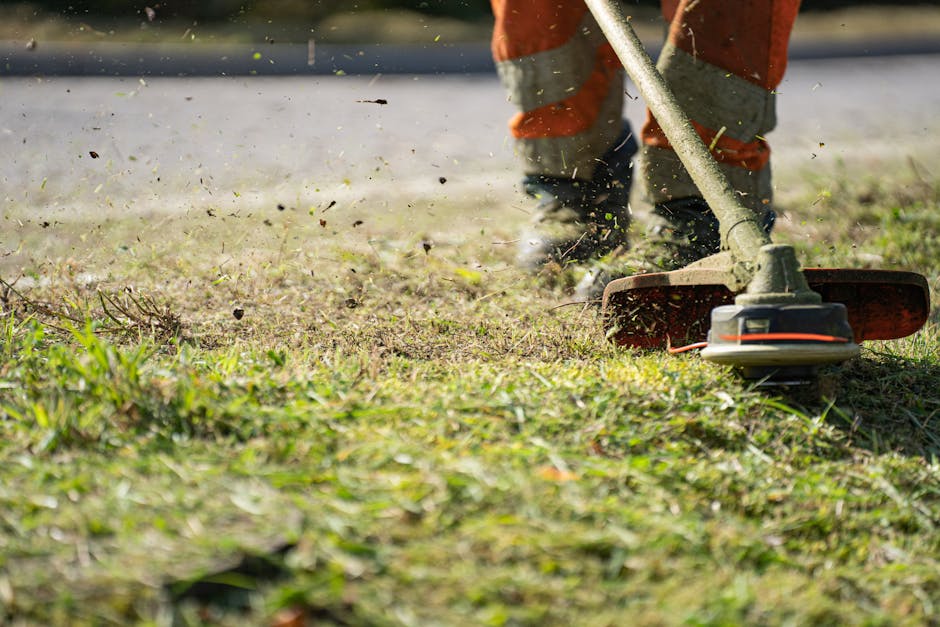 The image shows a gardener using a petrol-powered grass trimmer to trim the edges of a lawn in a garden. The gardener is wearing orange work trousers with grey reflective strips and sturdy shoes. The 