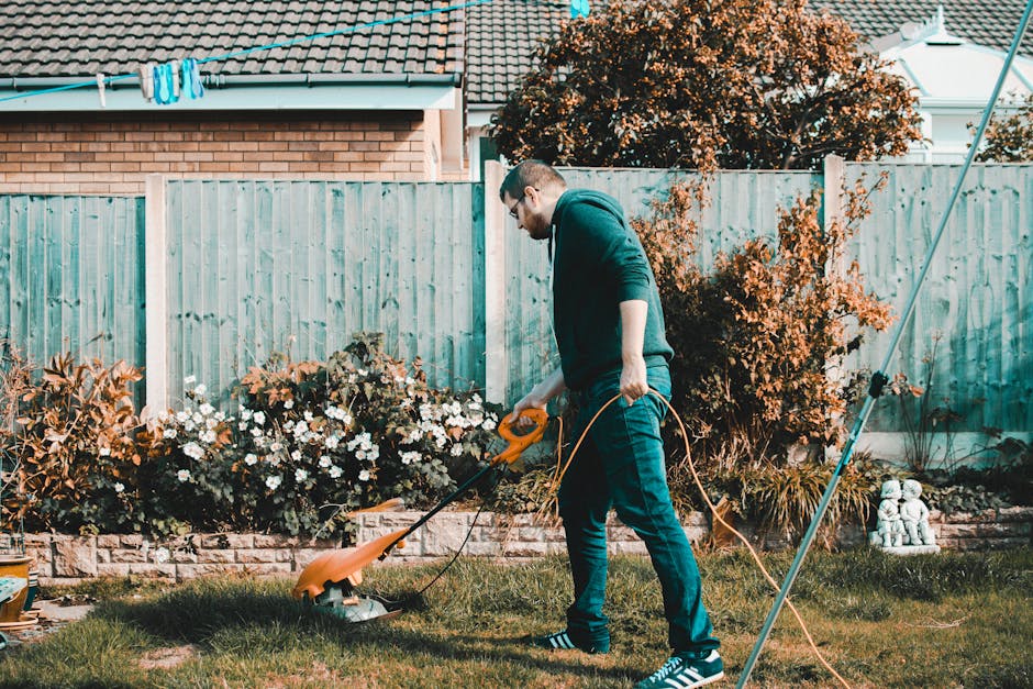 The image shows a gardener using a petrol-powered grass trimmer to trim the edges of a lawn in a garden. The gardener is wearing orange work trousers with grey reflective strips and sturdy shoes. The 