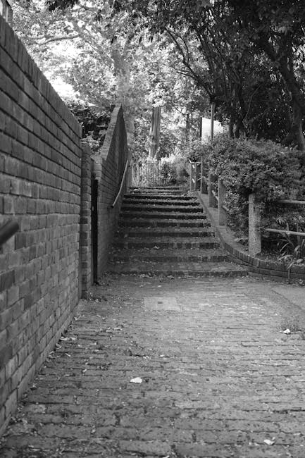 A black and white image of a narrow outdoor staircase in a residential garden setting, descending from a paved pathway made of uneven brick or stone. On the left side, a high brick wall runs parallel
